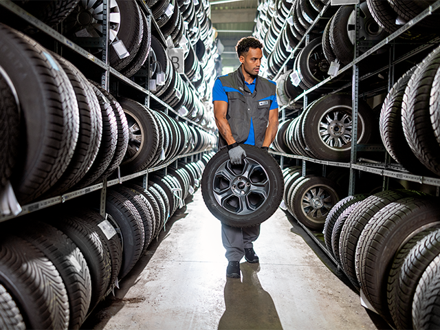 Mann in einem Lagerhaus trägt einen Reifen zwischen Regalen voller Reifen.