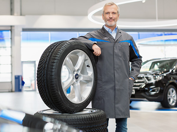 Mann in grauem Arbeitsmantel steht neben Reifen im Autohaus hinter einem Auto.