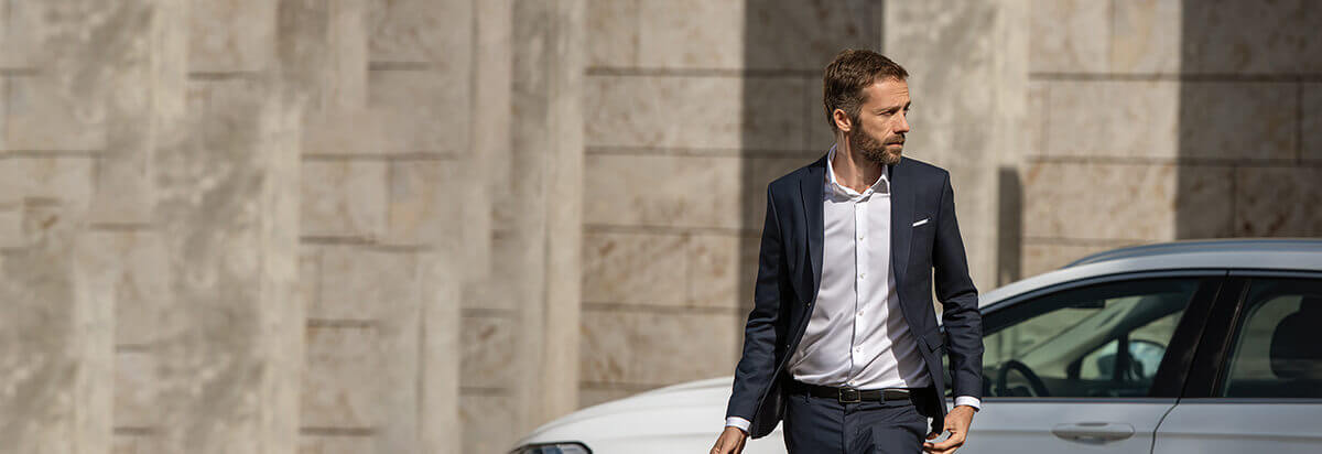 Ein Mann in einem Anzug geht an einem Auto vorbei, mit einem modernen Gebäudewand im Hintergrund.