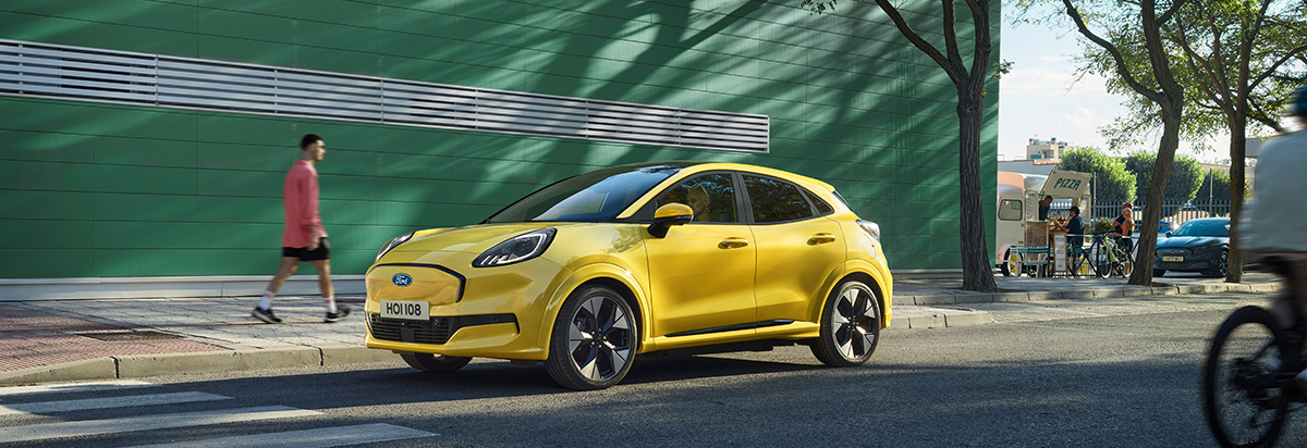 Gelber Ford Pkw auf einer Stadtstraße, im Hintergrund Passanten und ein food truck, grüne Wand.