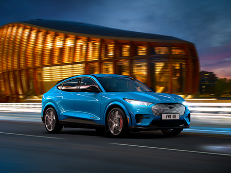 Ein blauer Ford Mustang Mach-E fährt bei Nacht vor einem modernen Gebäude entlang.