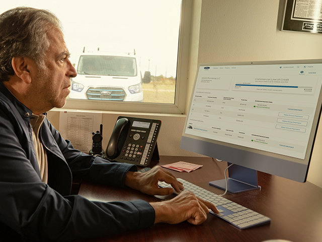 Mann arbeitet am Computer in einem Büro, Sicht auf eine Lieferung im Hintergrund. Bildschirm zeigt Kreditantrag.