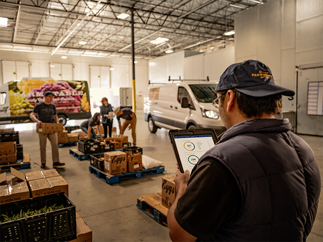 Mann mit Tablet in einem Lager, während andere Mitarbeiter Produkte in Kisten sortieren.