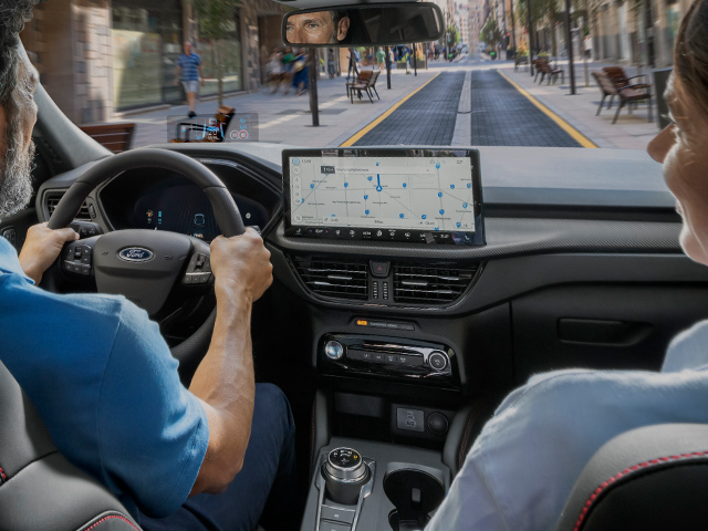Inneraum eines Autos mit einem Fahrer und Beifahrer, Bildschirm zeigt eine Karte, moderne Stadtstraße im Hintergrund.