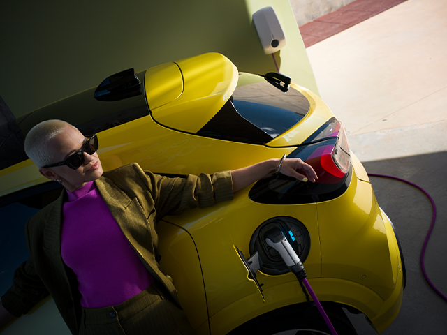 Frau mit kurzer Frisur und Sonnenbrille lädt ein gelbes Elektroauto an einer Ladestation auf.