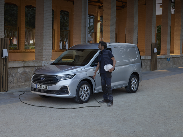 Ein silbernes Elektrofahrzeug wird aufgeladen, während ein Mann mit einem Helm daneben steht.