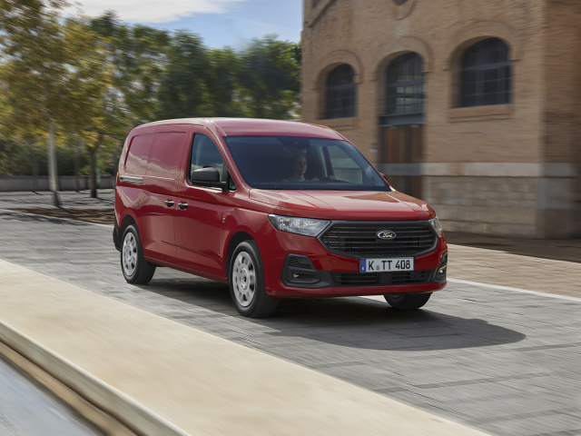 Ein roter Lieferwagen von Ford fährt auf einer gepflasterten Straße mit Bäumen im Hintergrund.
